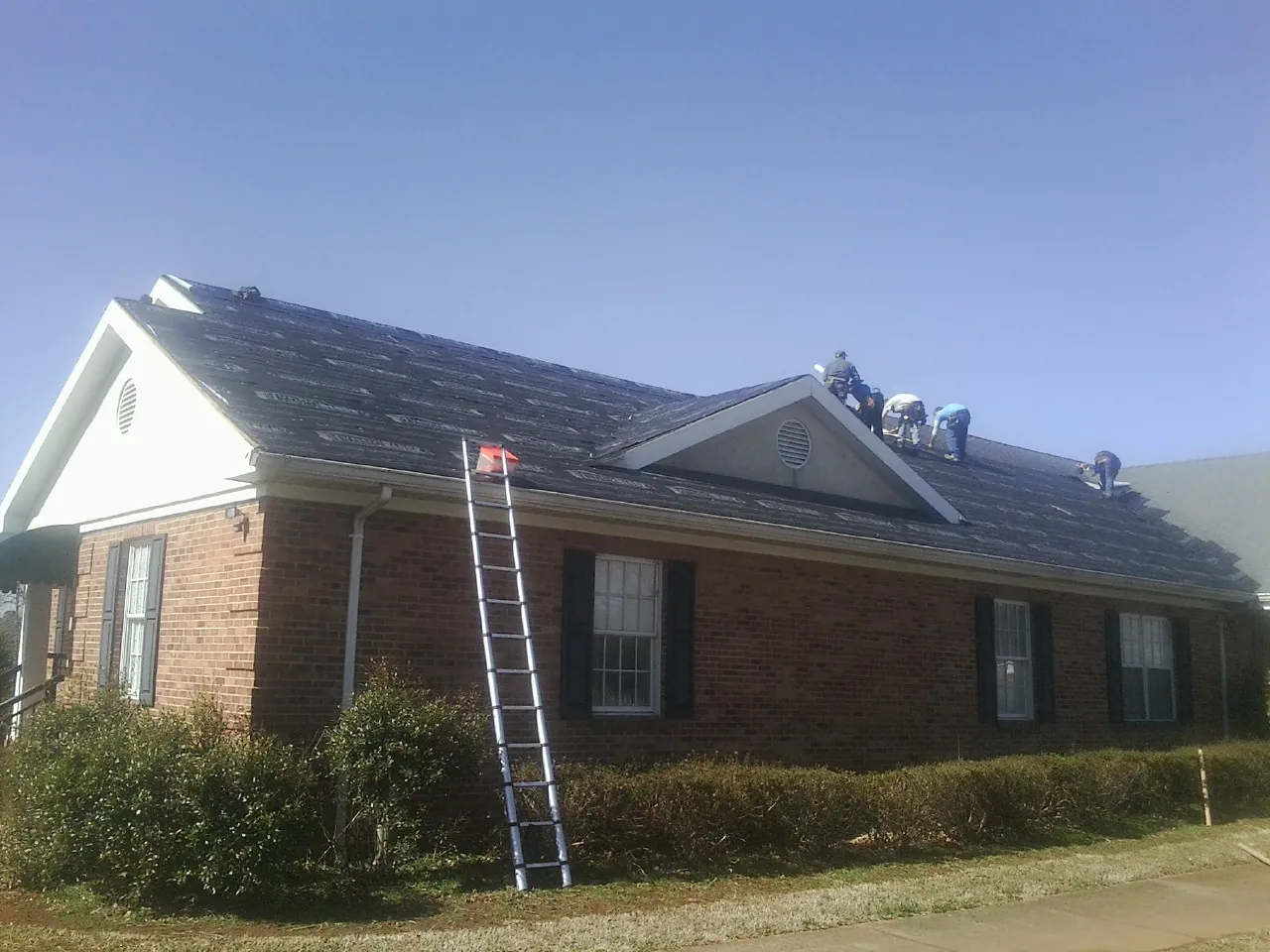 Crew tearing shingles off a brick ranch roof with a ladder set — Emergency Roof Repair work by our crew in Drayden