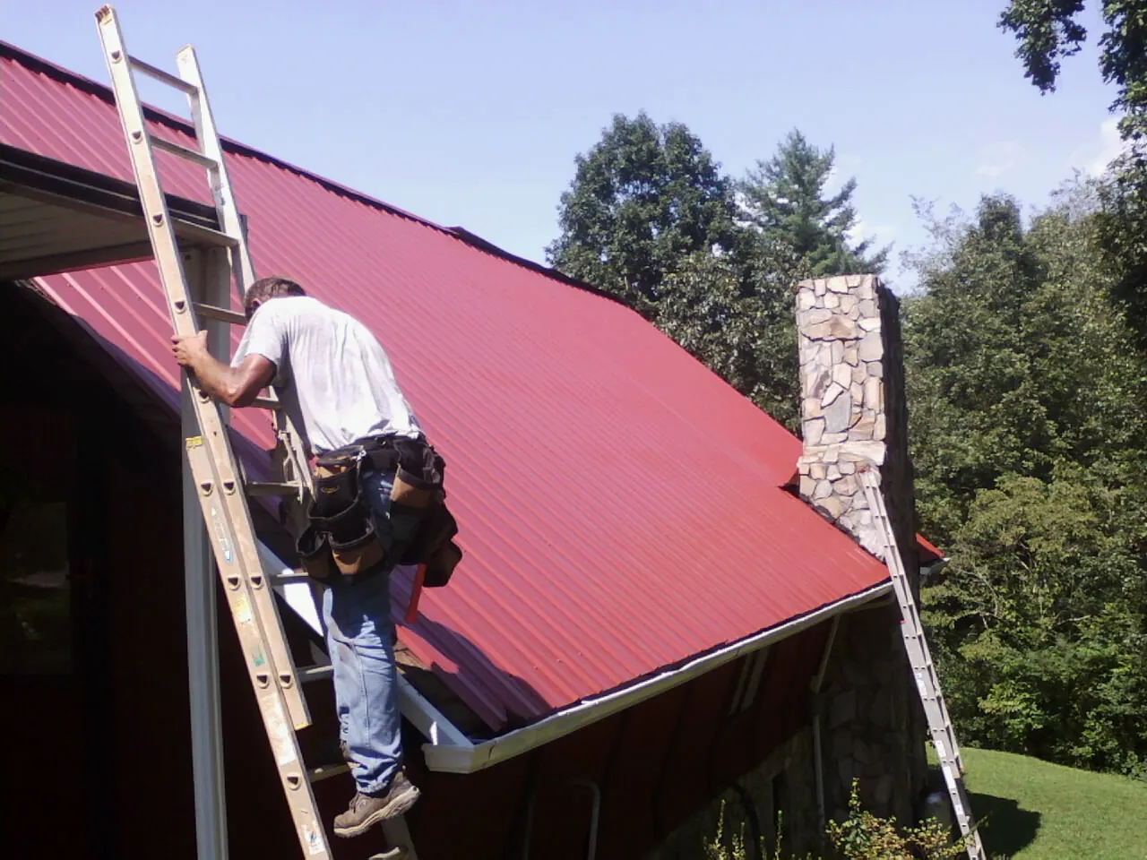 Roofer on a ladder beside a red metal roof and stone chimney — Soffit & Fascia Repair by Roofing Brothers in Drayden